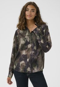 Donna con capelli castani mossi che indossa una camicia a maniche corte con motivo mimetico verde, marrone e nero e jeans scuri, su uno sfondo chiaro e semplice.