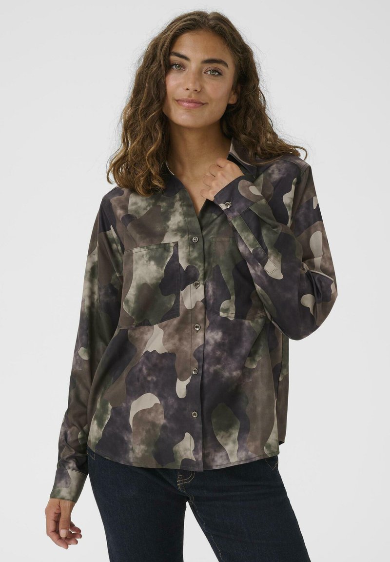 Donna con capelli castani mossi che indossa una camicia a maniche corte con motivo mimetico verde, marrone e nero e jeans scuri, su uno sfondo chiaro e semplice.