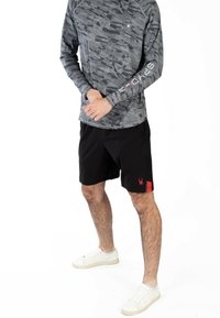 Maglietta a maniche lunghe in camo grigia con collo tondo, abbinata a pantaloni corti neri con un logo di ragno rosso, indossata con sneakers bianche.