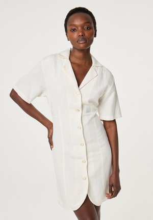 Vrouw draagt een wit overhemdjurk met korte mouwen, knopen en een kraag, staand met één hand op haar heup tegen een effen achtergrond.