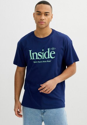 Joven con camiseta azul marino con texto verde "Inside" y la frase "Where Passion Meets Work", combinada con vaqueros azul claro, fondo neutro.
