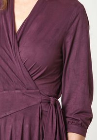 Lila Wickelkleid mit langen Ärmeln, aus weichem Stoff gefertigt. Mit einem Knoten-Detail an der Taille und einem tiefen V-Ausschnitt. Glatte Textur.