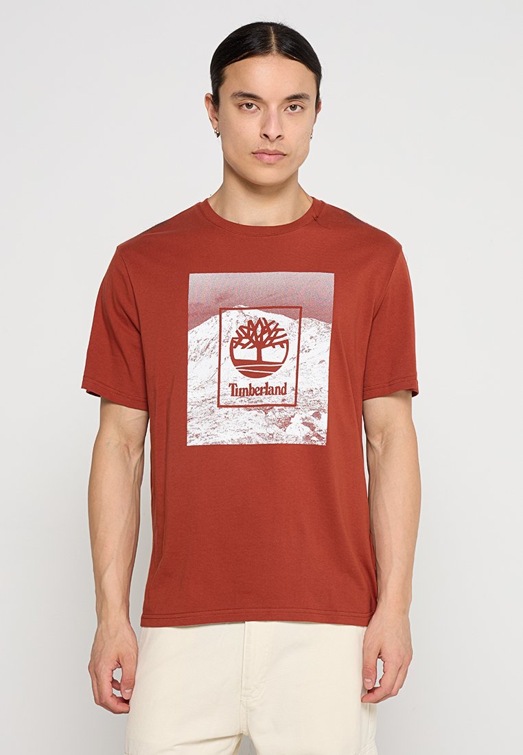 T-shirt in cotone rosso con collo a giro. La grafica centrata presenta un logo ad albero e la scritta "Timberland" su uno sfondo chiaro con montagne.