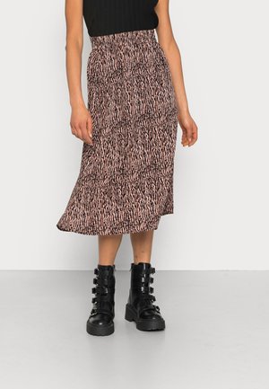 Femme portant une jupe midi rose et noire à motifs, un top noir sans manches et des bottes de combat noires à boucles, debout sur un fond clair.