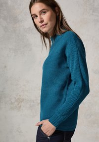 Türkisfarbener Strickpullover mit strukturiertem Rippmuster, hohem Kragen und langen Ärmeln, kombiniert mit dunklen Jeans. Dezenter Glanz auf dem Stoff.