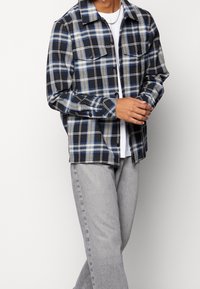 Koszula flanelowa w kratę w kolorze granatowym, niebieskim i beżowym. Długie rękawy, zapinana na guziki z dwiema kieszeniami na piersi, noszona na białym t-shircie. Szare dżinsy.