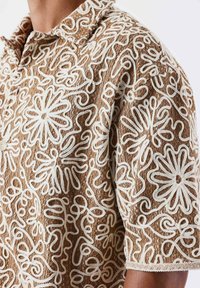 Camisa marrón de manga corta con una tela texturizada que presenta patrones de bordado florales y abstractos en blanco. Botonera en el cuello.