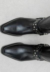 Botas de cuero negras hasta el tobillo con punta afilada, con una amplia correa decorada con tachuelas plateadas y un detalle de hebilla. Textura suave.