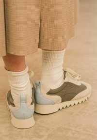 Le sneakers presentano un mix di materiali grigi, crema e azzurri, con un tessuto testurizzato e una suola bold e seghettata. Calze bianche a coste visibili.