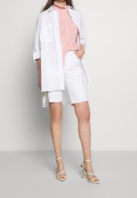 Woman wearing light pink buttoned blouse, long white open shirt, white knee-length shorts, and white high-heeled sandals standing against plain wall.