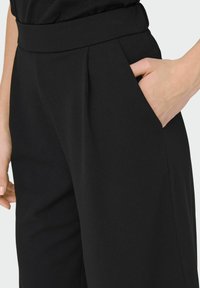 Svarta byxor med bred midja, utrustade med sidfickor och subtila veck framtill, tillverkade av ett slätt tyg med matt yta.