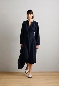 Robe rayée marine à manches longues et ceinture à la taille, agrémentée de deux poches avant ; portée avec des chaussures légères à enfiler.