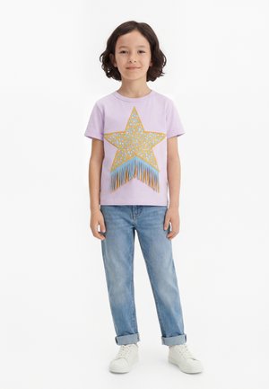 Otrok s temnimi lasmi nosi sivkasto majico z veliko rumeno zvezdo in modrimi resicami, svetle zavihane kavbojke in bele superge.