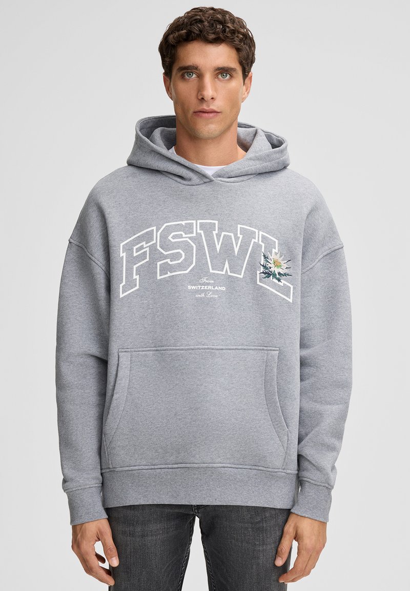 Uomo che indossa un felpa grigia con grandi lettere "FSWL" e un piccolo disegno di fiore, in piedi contro uno sfondo chiaro e semplice.