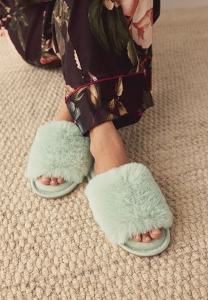 Füße, die mintgrüne, flauschige Hausschuhe tragen, kombiniert mit dunkelblumigen Schlafanzughosen auf einem beigen, strukturierten Teppich.