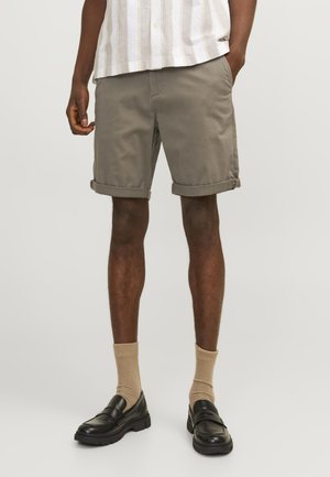Pantalones cortos de algodón caqui con puños doblados, combinados con calcetines beige y mocasines negros. El modelo está de pie frente a un fondo neutro.