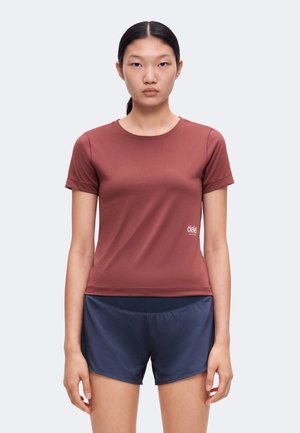 Ung kvinde med mørkt hår i en hestehale iført en tætsiddende brun kortærmet skjorte og navyblå sportsshorts, stående mod en ensfarvet baggrund.