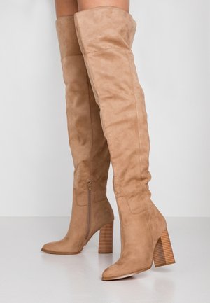Bottes beiges au-dessus du genou en daim, avec un talon épais empilé, un détail de fermeture éclair sur le côté et une texture lisse sur l'ensemble.
