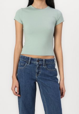 Kvinde iført en tætsiddende lys grøn kortærmet crop top og højtaljede blå denimjeans, stående mod en ensfarvet baggrund.