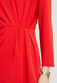 Rød kjole med lange ærmer, der har lodrette plisseringer og en figursyet talje. Glat tekstur og et guldur på håndleddet tilføjer detaljer.