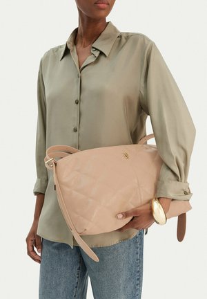 Persona con camisa abotonada color verde oliva y jeans azules sosteniendo un bolso grande acolchado beige con detalles en dorado.