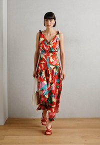 Robe maxi colorée et à motifs avec un décolleté en V, jupe à volants et liens aux épaules ; portée avec des sandales à lanières rouges et un petit sac à main blanc.