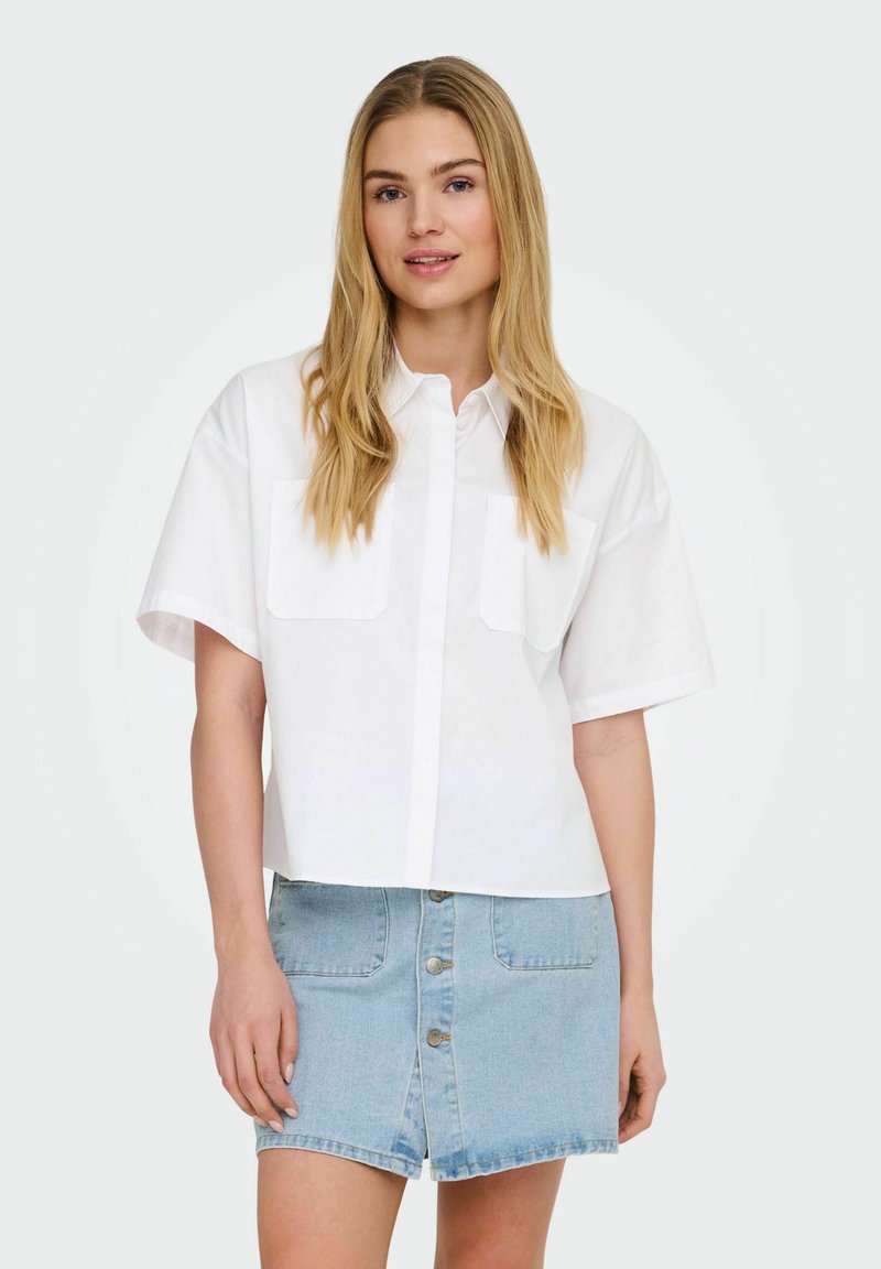 Jeune femme aux longs cheveux blonds portant une chemise blanche à manches courtes boutonnée et une jupe en jean bleu clair avec des boutons à l'avant.