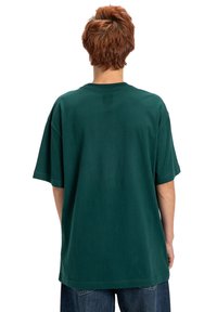 Zielona oversize'owa koszulka, wykonana z bawełny, krótki rękaw, okrągły dekolt, gładki tył z minimalnymi detalami, w połączeniu z luźnymi niebieskimi dżinsami.