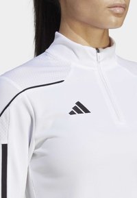 Weißes Sportoberteil mit einem Viertel-Reißverschluss-Kragen, strukturierten Schulterakzenten und schwarzen Streifen. Enthält ein unauffälliges schwarzes Logo auf der Brust.