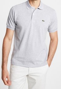 Polo gris clair en tissu mélangé coton, avec patte de boutonnage à deux boutons, manches courtes, et petit logo Lacoste vert sur la poitrine.