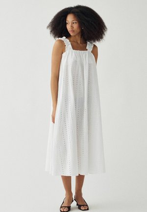 Žena s objemnými vlasmi má na sebe biele midi šaty bez rukávov s dierkovaným vzorom, volánikovými ramienkami a čiernymi otvorenými lodičkami, stojaca pri obyčajnej stene.