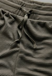 Közeli kép szürke, gofrös kötésű, húzózsinórral ellátott derékrészű nadrágról, melynek anyagából készült zsinórja megkötve, és texturált anyag részletei láthatók.
