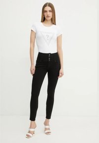 T-shirt blanc ajusté avec un graphique triangulaire gris, associé à un jean skinny noir taille haute avec fermeture à boutons et poches. Sandales à talons blanches.