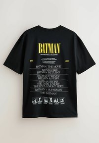 Čierne tričko s krátkym rukávom so bielym textom uvádzajúcim Batmanove filmy od roku 1966 do 2022, s veľkým žltým logom "BATMAN" navrchu.