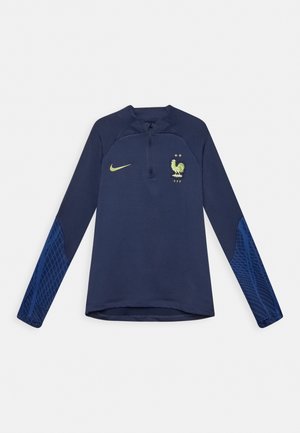 Granatowa, długa koszulka sportowa z rękawami, z zamkiem błyskawicznym do jednej czwartej, żółte logo Nike, emblemat drużyny piłkarskiej Francji oraz wzorzysta tkanina na dolnych rękawach.