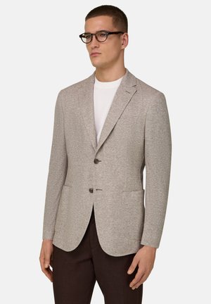 Man met bril in schildpadlook, beige gestructureerd colbert met twee knopen over een wit overhemd, en donkerbruine broek tegen een effen achtergrond.
