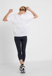 Vrouw in zwarte leggings en een wit mesh-top die met opgetilde armen wegloopt tegen een effen witte achtergrond.