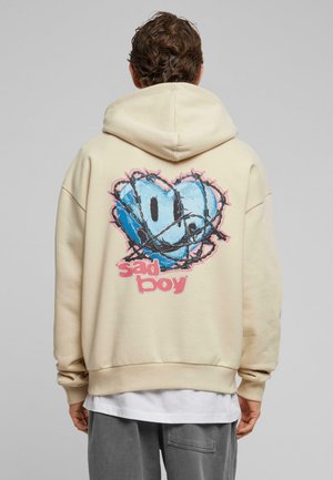 Beige hættetrøje med stort blåt hjertegrafik, detalje med pigtråd og "sad boy" tekst i pink. Den har afslappet pasform og en blød tekstur.