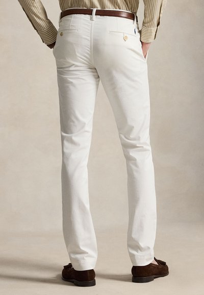 Άνδρας που φοράει λευκό παντελόνι slim-fit, καφέ ζώνη, ριγωτό μπεζ πουκάμισο μέσα από το παντελόνι και σκούρα καφέ μοκασίνια, στέκεται γυρισμένος.