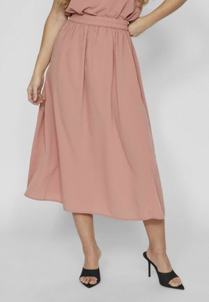 Vrouw draagt een vloeiende midi-rok in stoffig roze met een elastische tailleband, gecombineerd met zwarte open teen hoge hakken mules, staand tegen een grijze achtergrond.