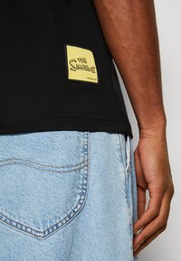 Crna pamučna majica s žutim logom "The Simpsons" na rubu, uparena s svijetloplavim traper šortsevima s zakrivljenim šavovima na džepovima.