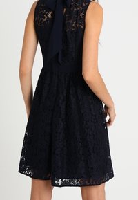 Ärmlös klänning i marinblå spets med A-linjeformad kjol, figursydd midja och en stor rosett knuten i nacken.