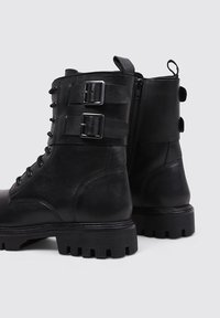 Bottes de combat en cuir noir avec un laçage à l'avant, deux sangles à boucle, une fermeture éclair sur le côté et une semelle en caoutchouc épaisse et texturée.