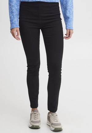 Zwarte high-waisted legging met een soepele textuur en een strakke pasvorm, gecombineerd met grijze en witte sneakers met groene accenten.