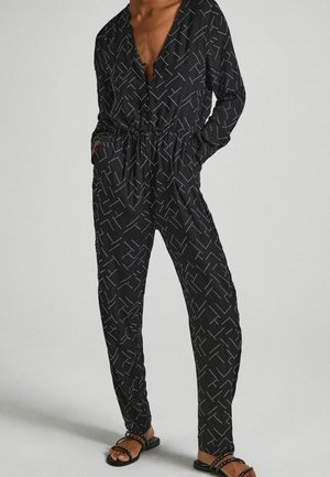 Kvinde iført en sort jumpsuit med hvide prikkede geometriske mønstre, lange ærmer, dyb V-hals, elastisk talje og sorte sandaler med nitter.