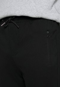 Svarta träningsbyxor med en slät yta, med dragsko i midjan och en ficka med blixtlås på sidan.