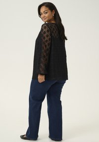 Top din dantelă neagră cu mâneci lungi și model floral, purtat cu blugi evazați de culoare albastru închis. Modelul stă cu o ușoară întoarcere, evidențiind ținuta.