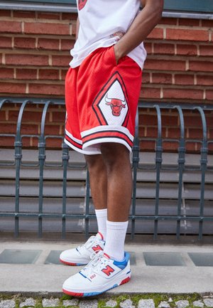 Persoon die rode Chicago Bulls basketbalshorts draagt, witte enkelsokken en witte New Balance sneakers met blauwe en rode accenten, staand in de buitenlucht.