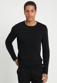 Pulover negru cu manșete lungi și guler la baza gâtului. Are un material texturat și o siluetă ajustată, asortat cu pantaloni închiși la culoare.