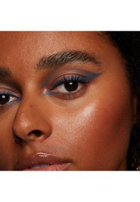Primo piano di un viso con eyeliner blu, labbra lucide e toni caldi della pelle. Sono visibili anche la pelle con texture e sopracciglia definite.